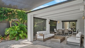 A modern outdoor patio with a white sofa, a wooden coffee table, and a dining area with wooden chairs and a table. - Spacious 4BR Poolside Escape (Burleigh Waters)