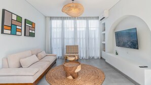 Ein modernes Wohnzimmer mit einer Couch, einem runden Holztisch, einer Hängeleuchte und einem großen Fenster mit durchsichtigen Vorhängen.