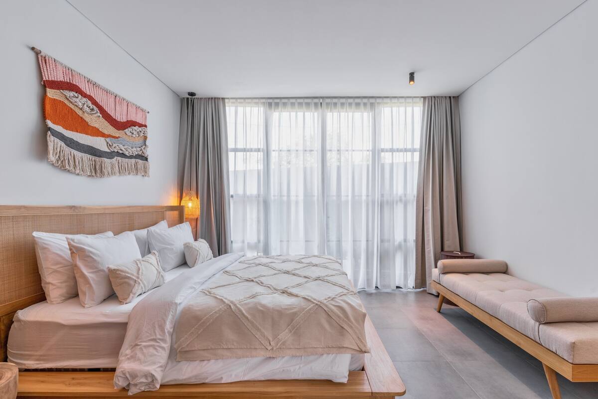 Quarto moderno com uma cama, um banquinho e uma janela grande.