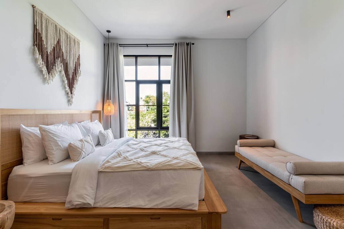 Quarto com cama, mesa de cabeceira, banqueta e uma janela grande com cortinas.