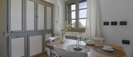 Comedor con mesa de madera, sillas blancas, un florero con plantas secas y una ventana con cortinas.