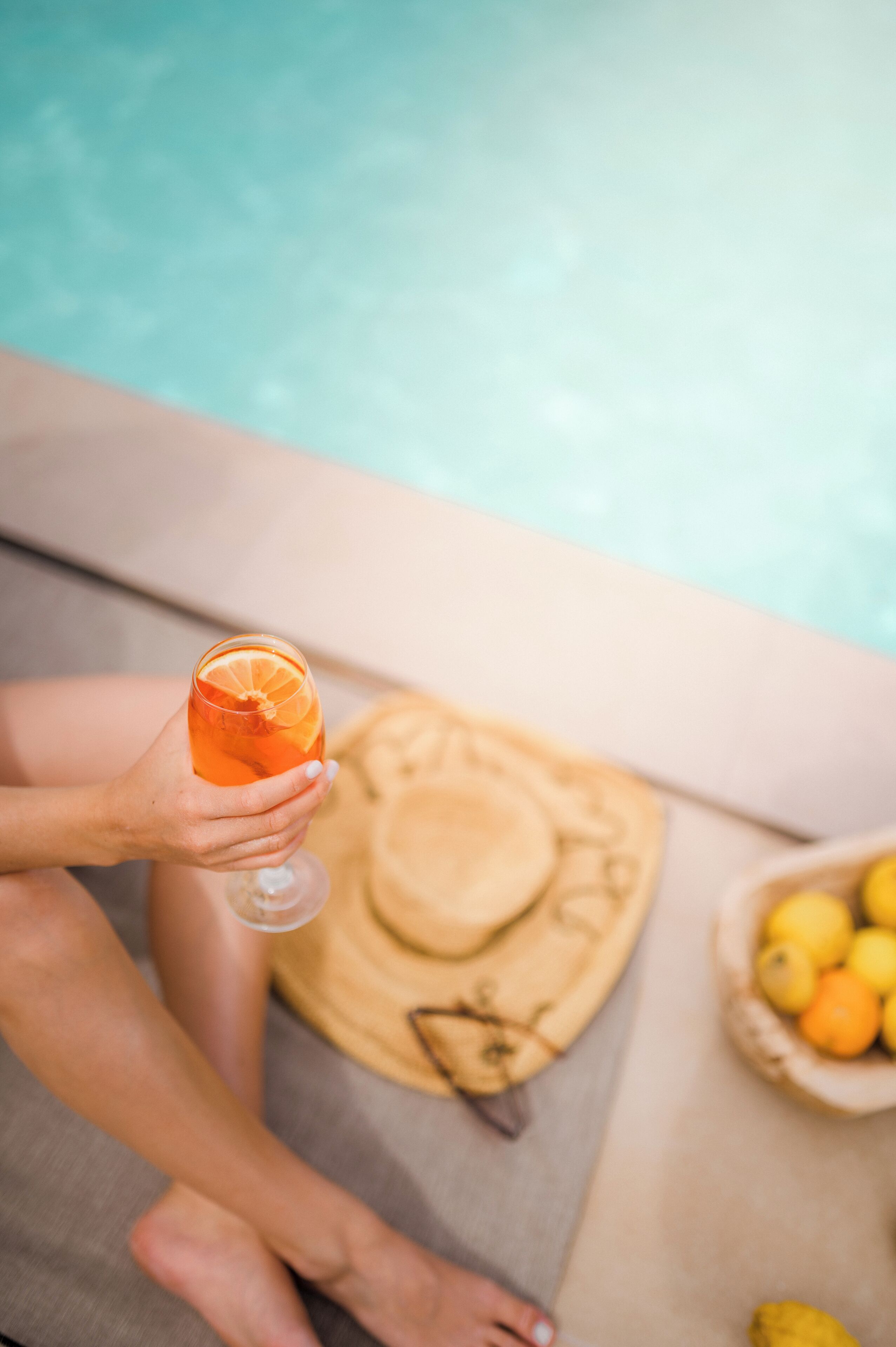 A person holding a drink with an orange slice, sitting by a pool with a hat and a bowl of lemons and oranges on the side.