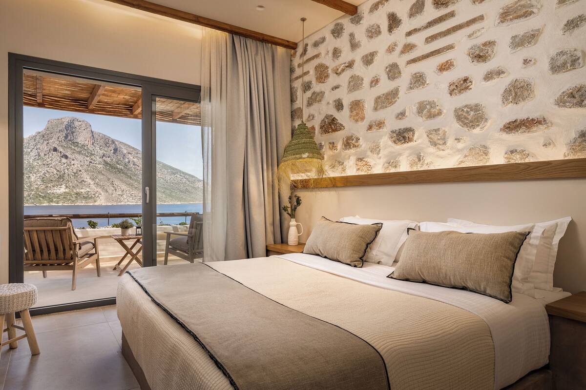 Un dormitorio con una cama grande, vista al mar, zona de comedor y pared de piedra.