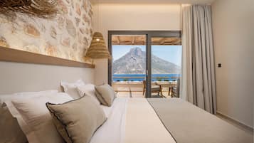 A bedroom with a large bed, a view of a mountain and sea, and a balcony with a table and chairs.