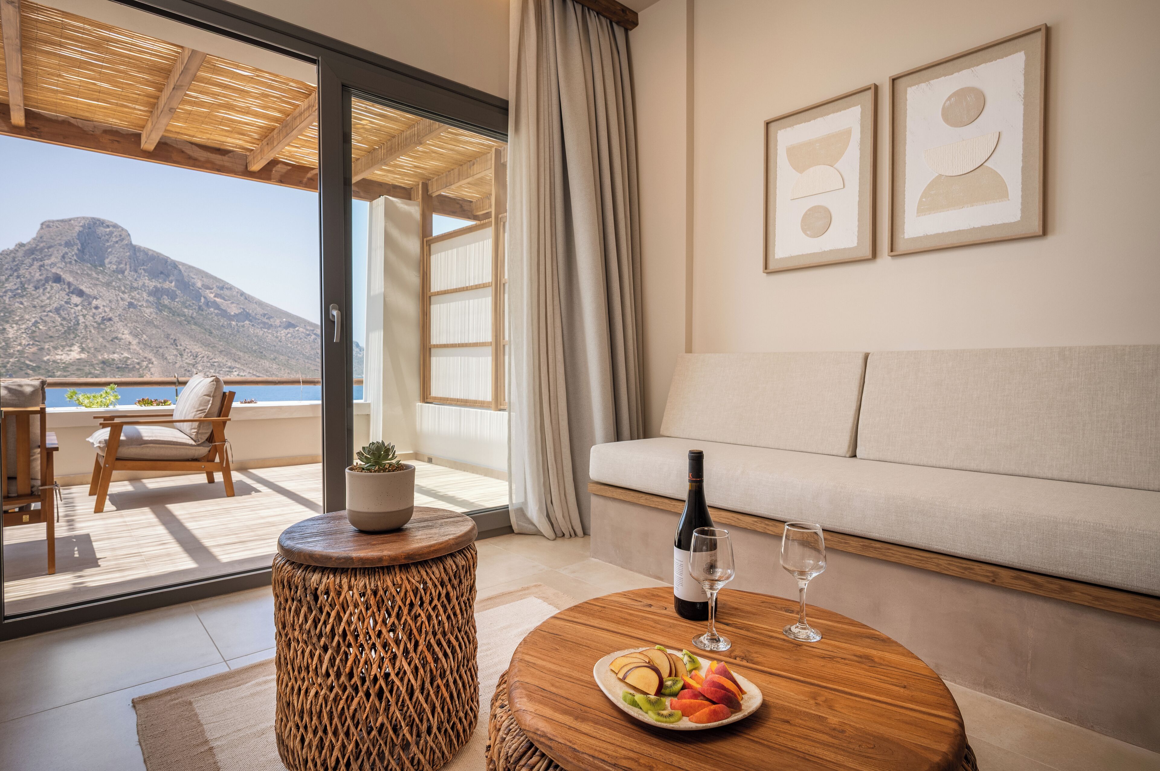 Una sala de estar moderna con un sofá, una mesa de comedor con un plato de fruta, una botella de vino y una copa. Hay un taburete de mimbre y una planta en maceta. La habitación tiene ventanas amplias con vistas a una montaña y al mar.