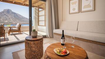 Una sala de estar moderna con un sofá, una mesa de comedor con un plato de fruta, una botella de vino y una copa. Hay un taburete de mimbre y una planta en maceta. La habitación tiene ventanas amplias con vistas a una montaña y al mar.