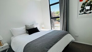A modern hotel room with a large bed, a nightstand, a lamp, and a window with a view of trees and sky. - New Merivale townhouse, centrally located by Hagley Park and City. (Christchurch)