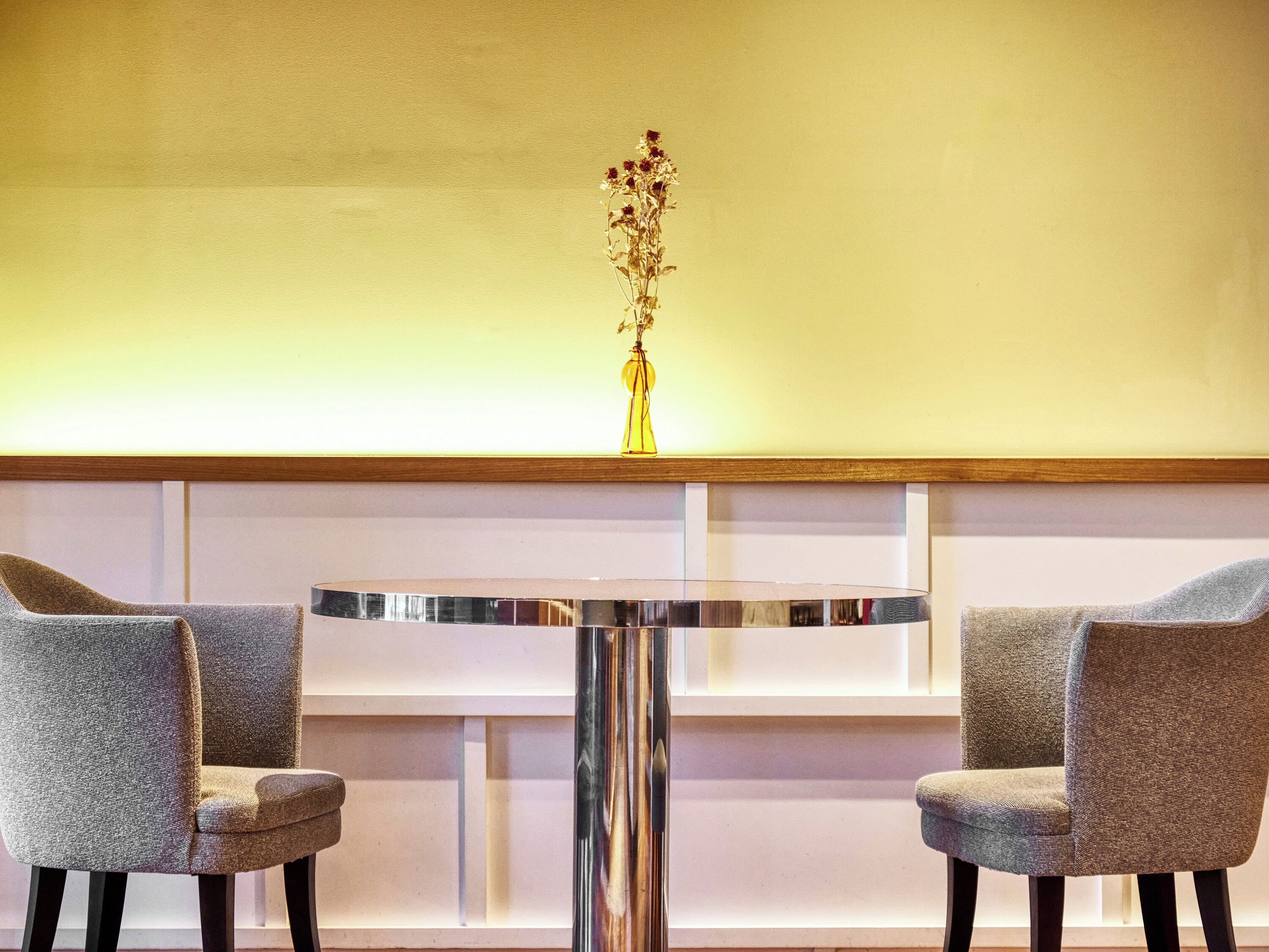 A round table with two grey upholstered chairs, a vase with flowers on the table, and a wooden shelf above.