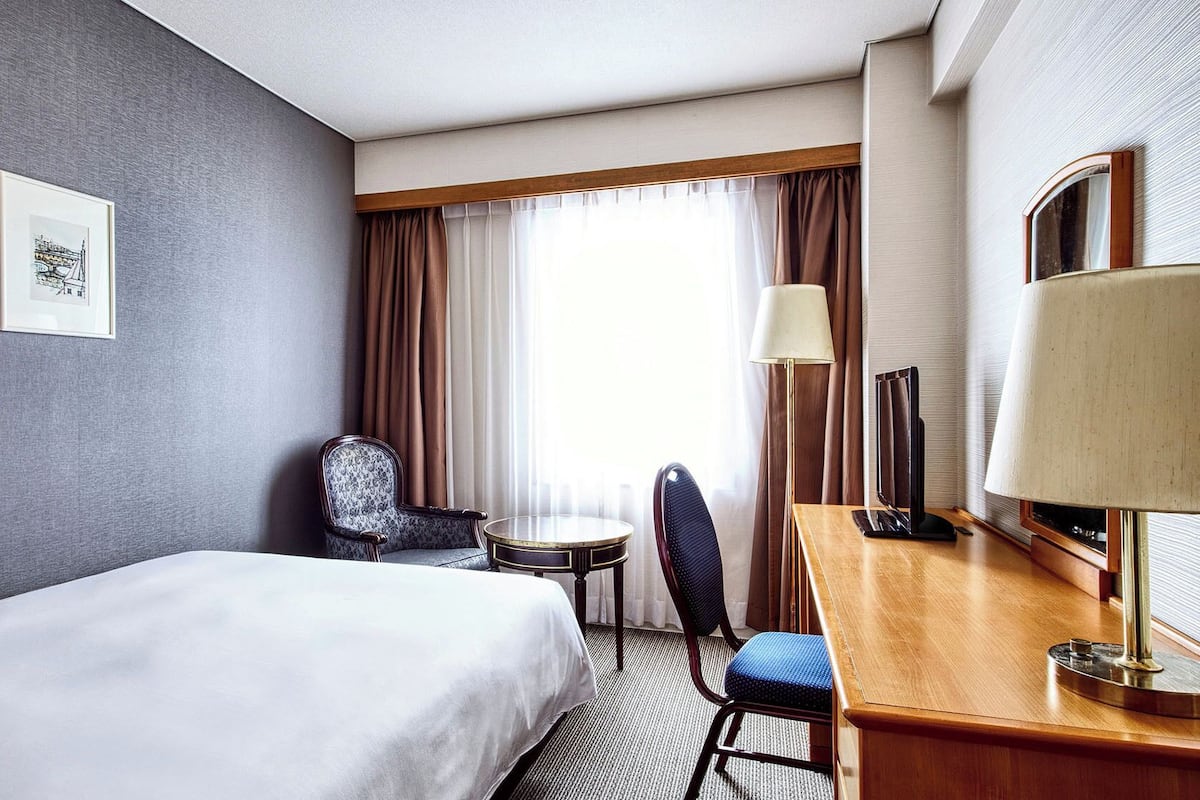 Une chambre d’hôtel avec un lit, un bureau, une chaise, une lampe, un miroir et une fenêtre avec des rideaux.
