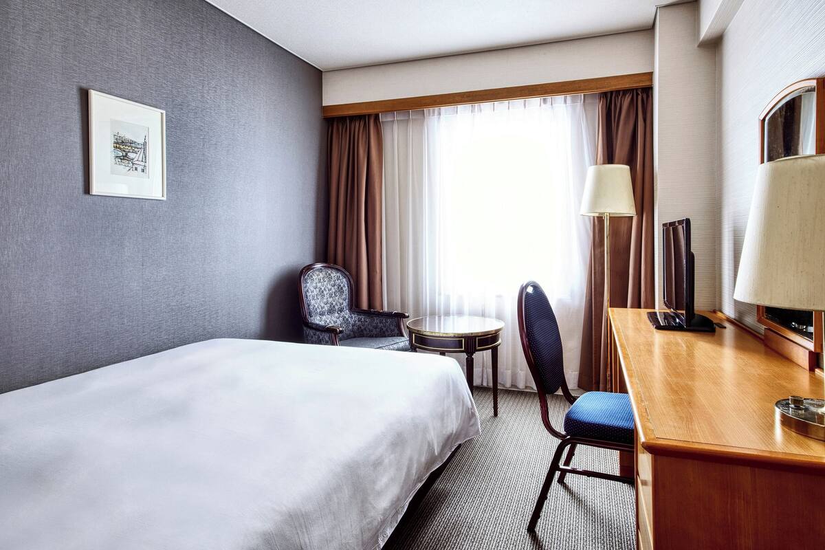 Une chambre d’hôtel avec un lit, un bureau doté d’une lampe, une chaise, une petite table et un tableau encadré au mur.