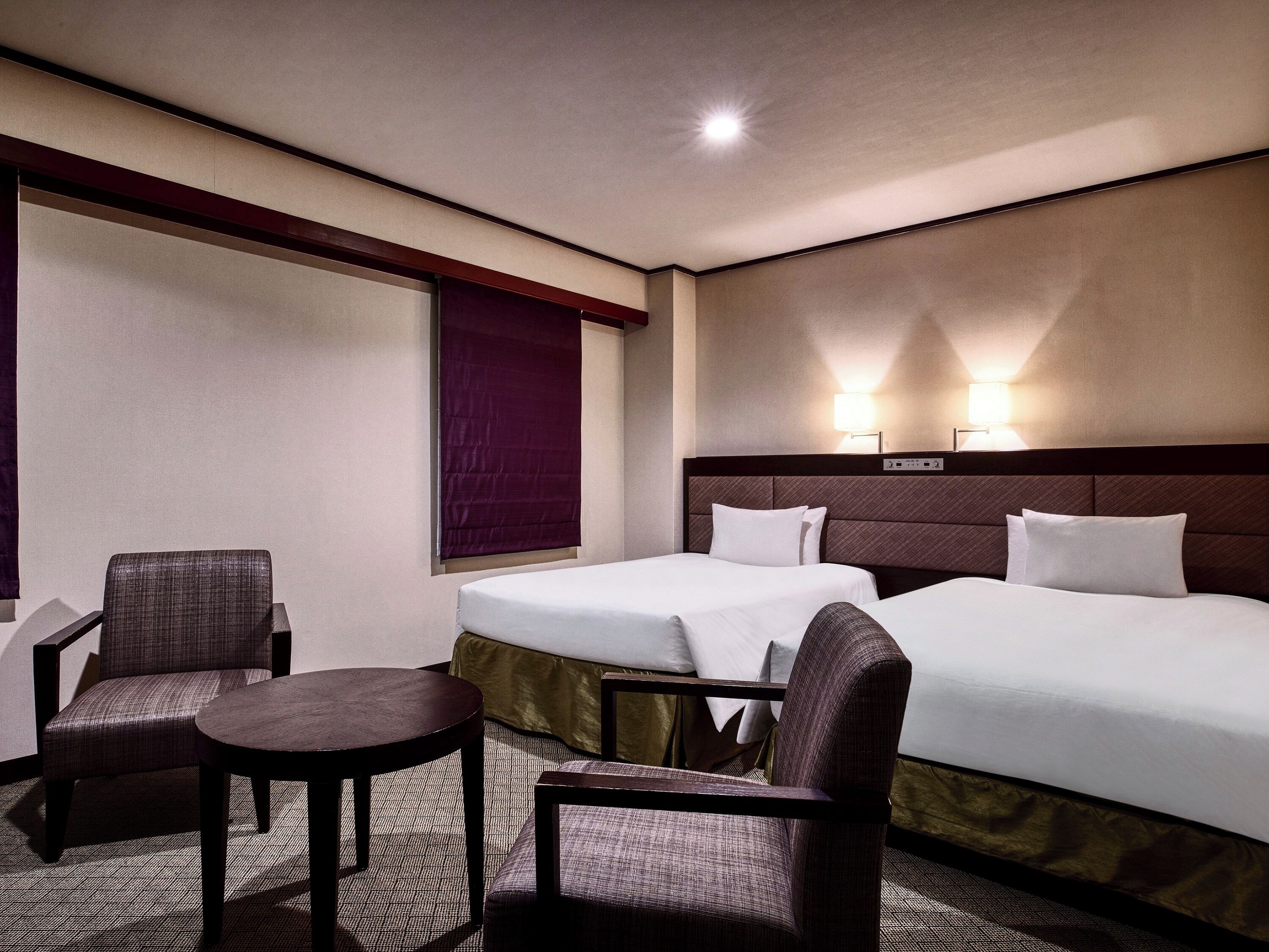 Hotelkamer met twee bedden, een klein tafeltje, twee stoelen, een raam met jaloezieën en een wandlamp.