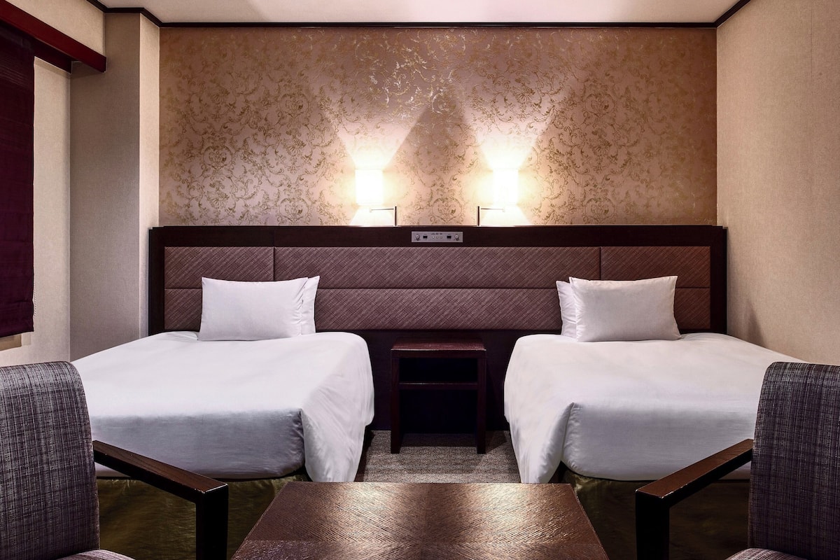 Une chambre d’hôtel avec deux lits, une table de chevet, une table et un luminaire fixé au mur.