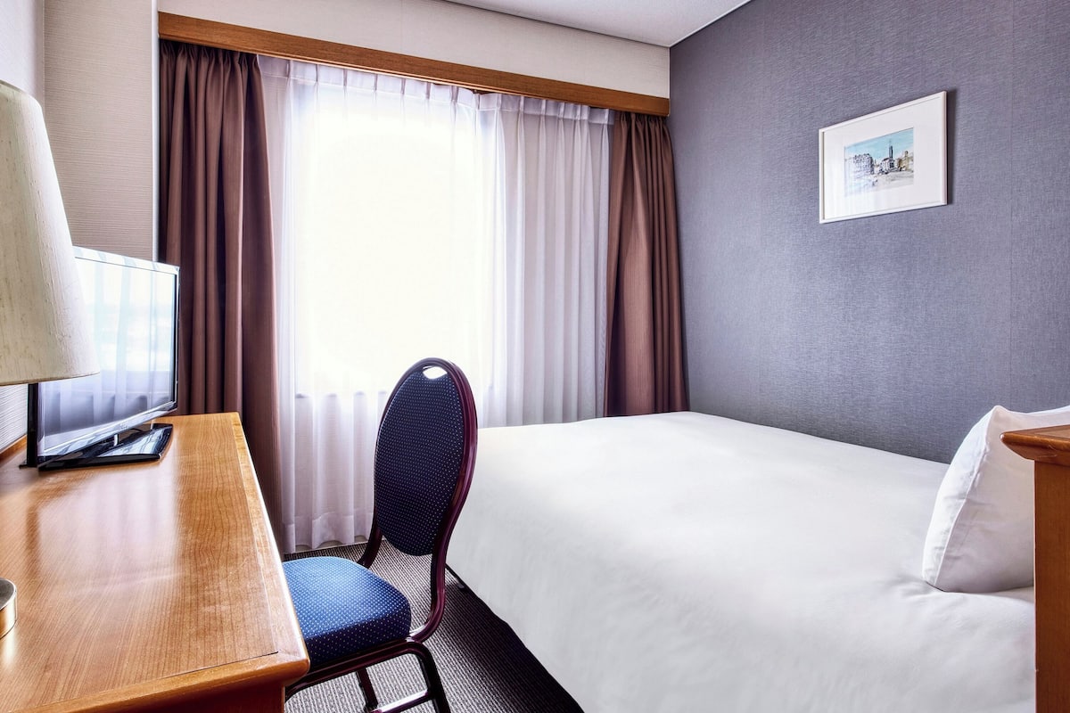 Une chambre d’hôtel avec un lit, un bureau avec une lampe, une chaise et une fenêtre avec des rideaux.