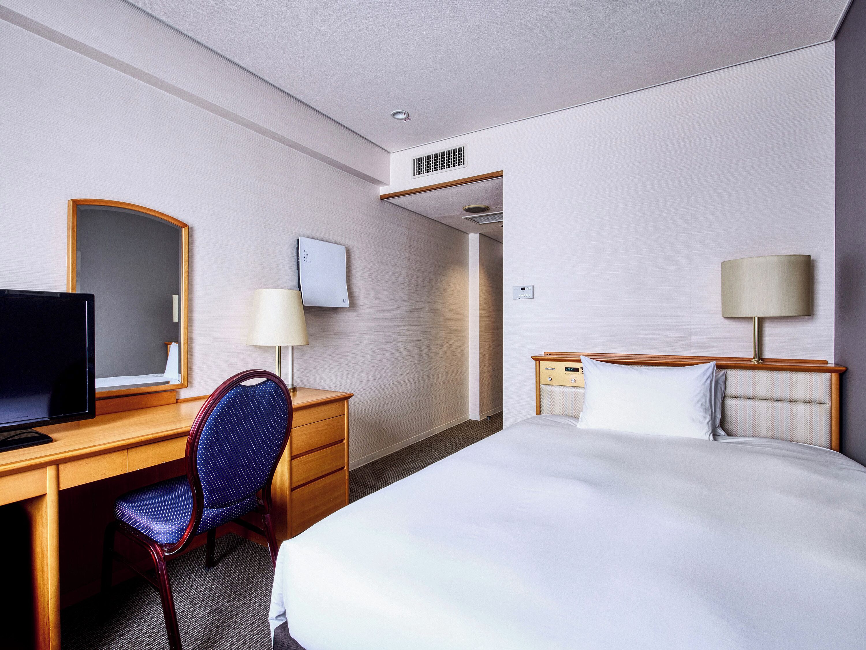 A hotel room with a bed, a desk with a computer, a chair, a TV, a lamp, and a bedside table.