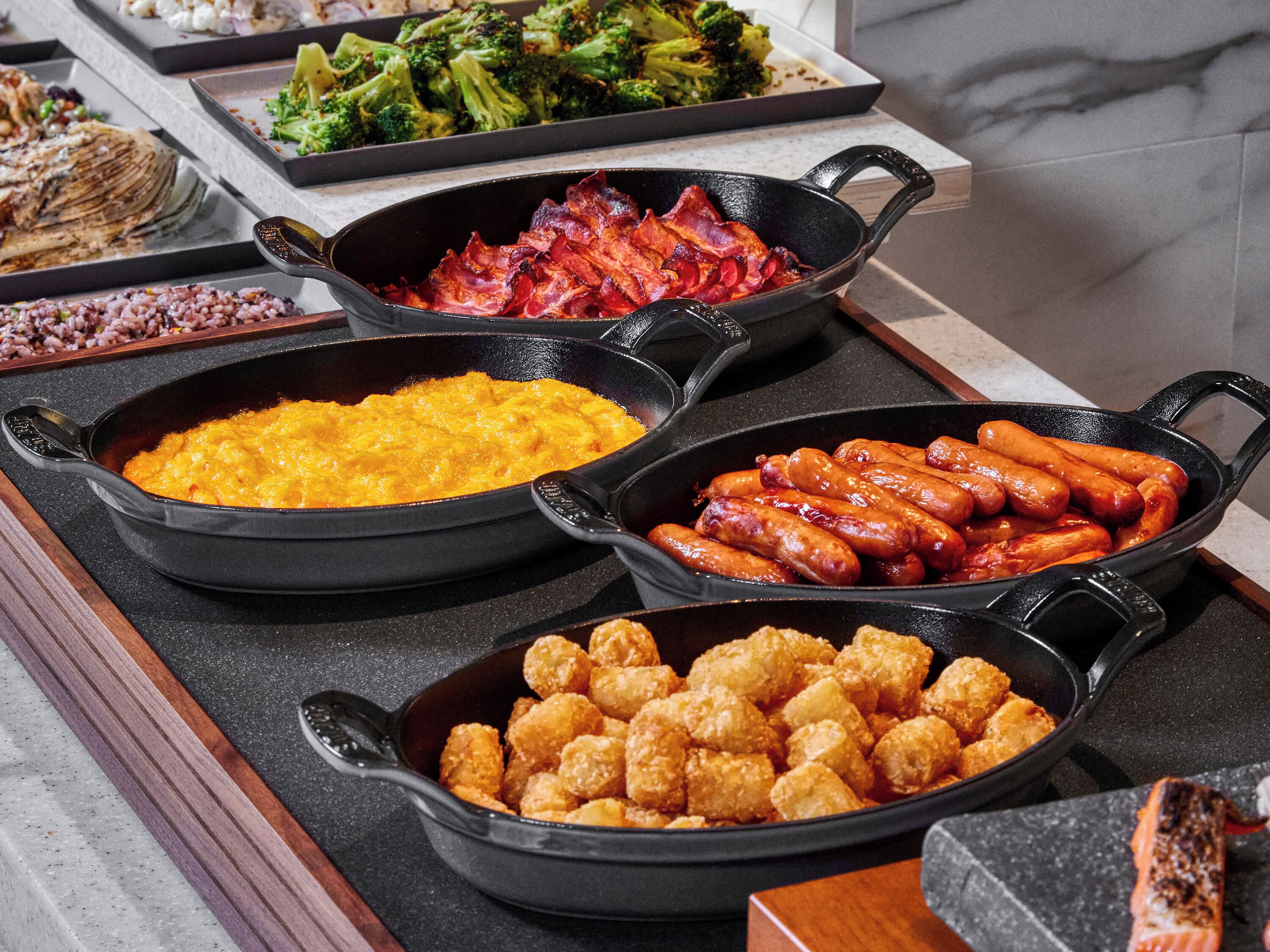 A buffet with various dishes including scrambled eggs, bacon, sausages, and tater tots.