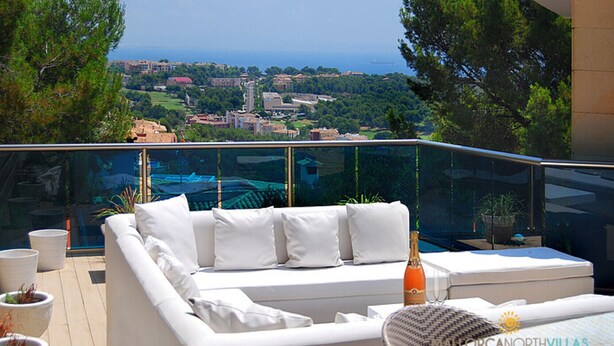 A balcony with a white sofa, a glass railing, and a view of a coastal town. - Villa Ocean View en Portals Nous by Mallorca North Villas (Portals Nous)
