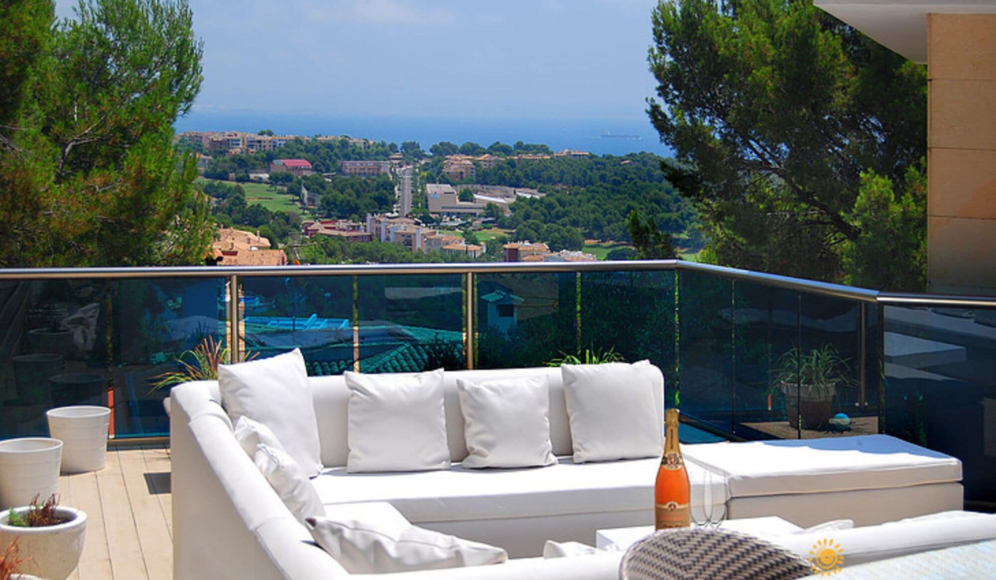A balcony with a white sofa, a glass railing, and a view of a coastal town.