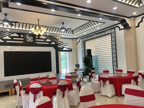 A conference room with round tables, white chairs with red sashes, a large screen, ceiling fans, and a modern ceiling design. - Bac Ninh Royal Hotel (Vo Cuong)