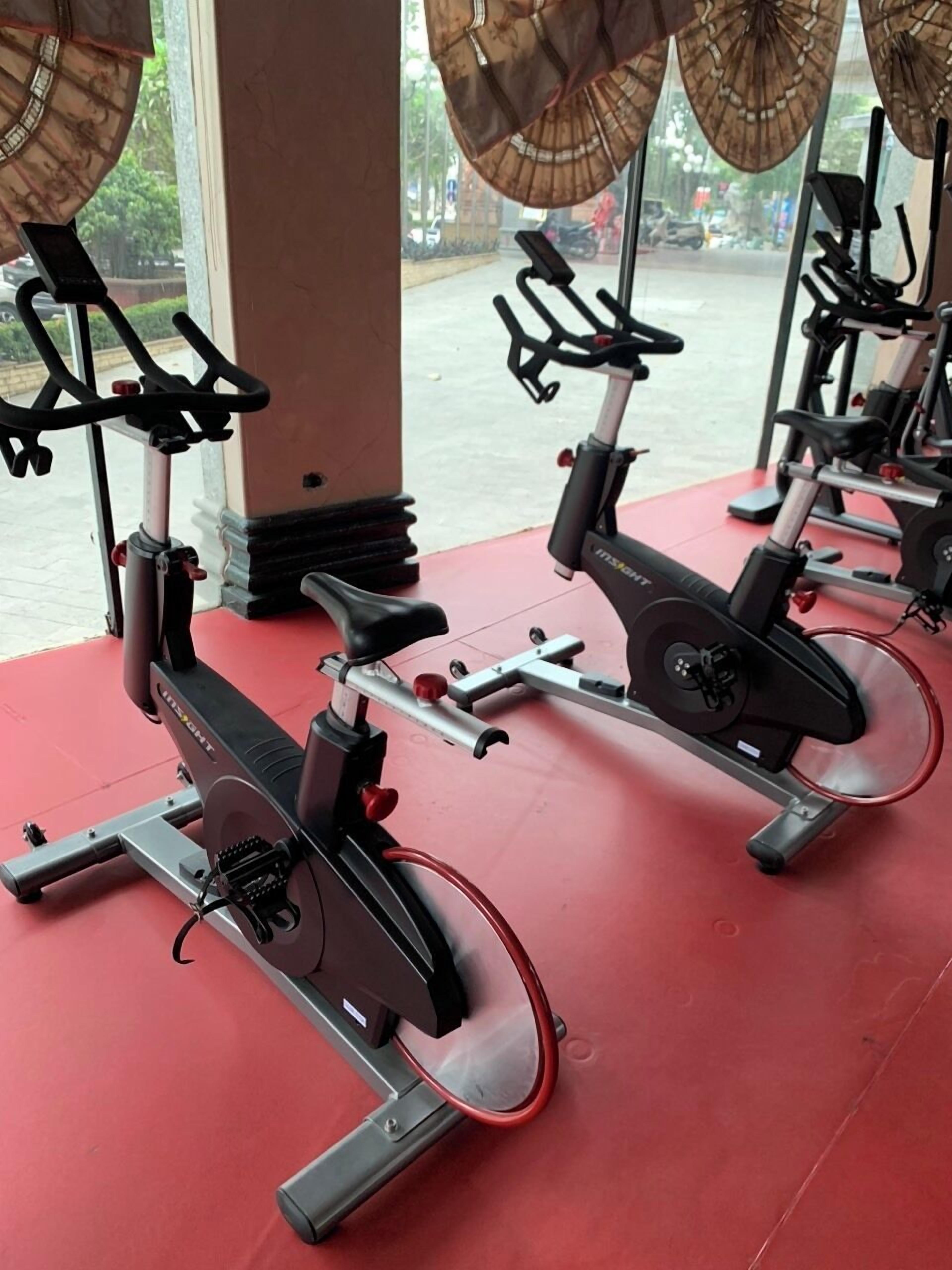Fileira de bicicletas ergométricas numa academia com piso vermelho e janelas amplas.