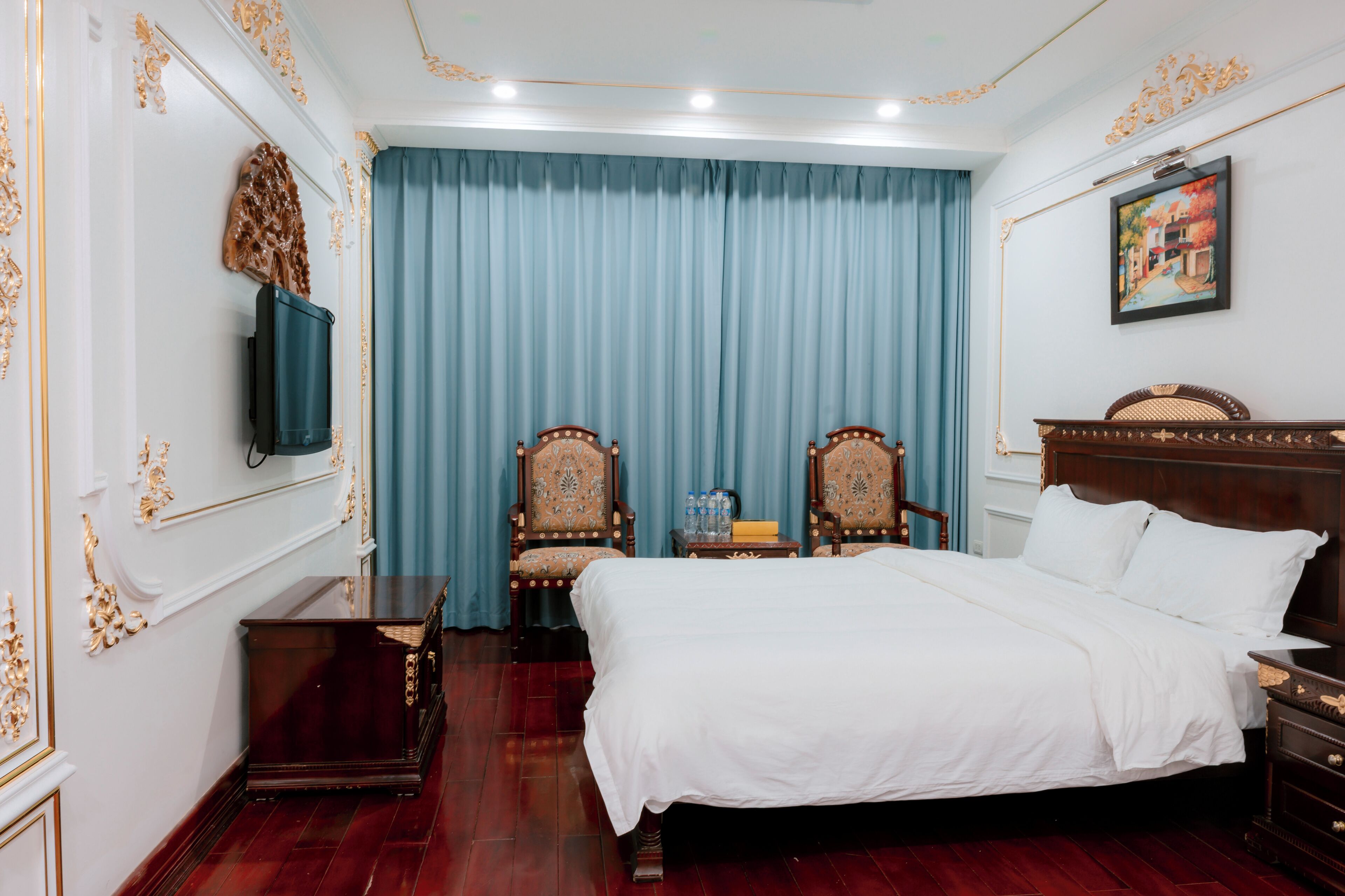 Une chambre à coucher avec un grand lit, deux fauteuils décoratifs, une petite table avec des bouteilles et un téléviseur fixé au mur.