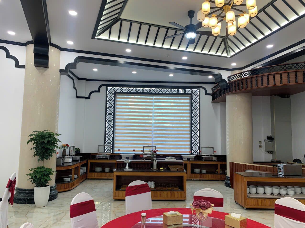Un amplio hall de hotel con una zona de estar central, un ventanal con persianas y un ventilador de techo.