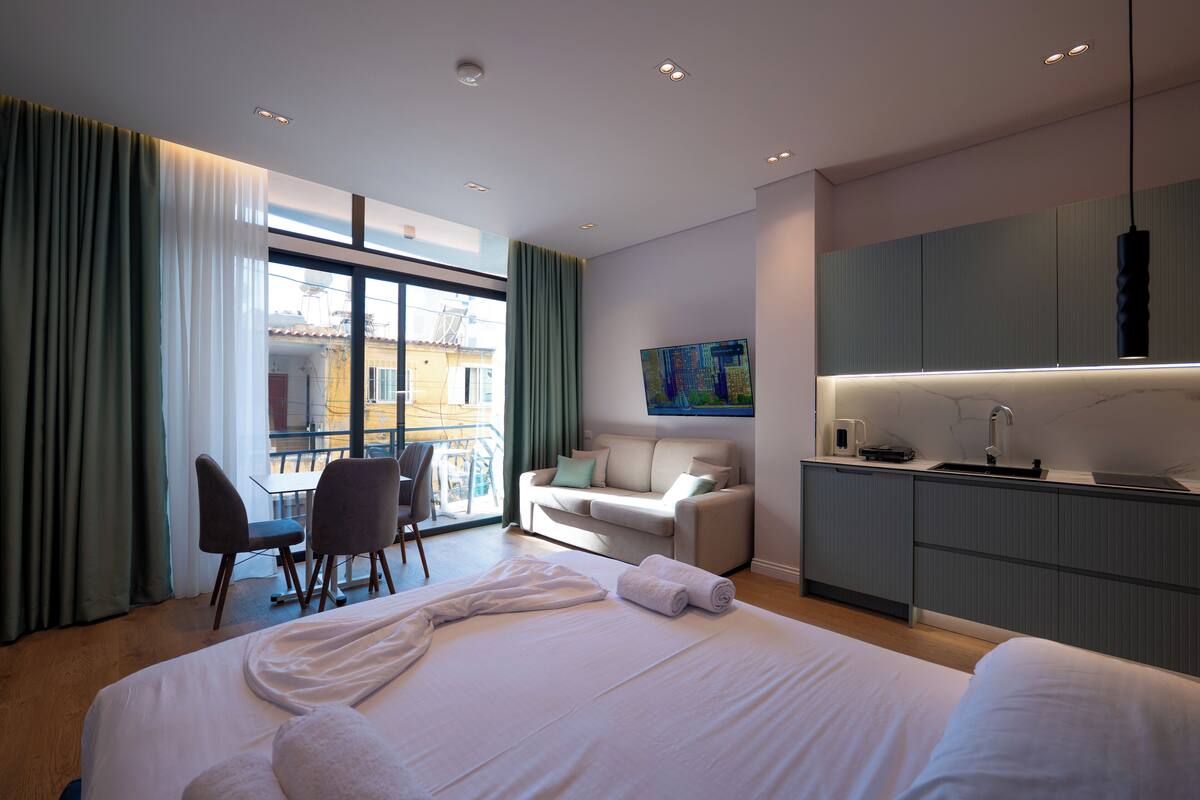 A modern hotel room with a large bed, a seating area with a sofa and chairs, a kitchenette, and a balcony with a view.
