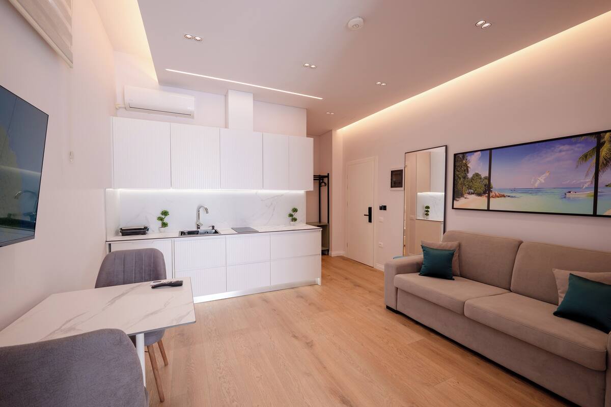 A modern living room with a sofa and a dining table, a kitchen area with white cabinets, and a large window with a view of the beach.