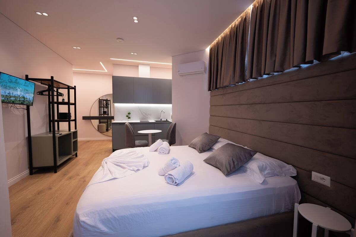 A modern hotel room with a large bed, a TV, a desk, and a bathroom in the background.