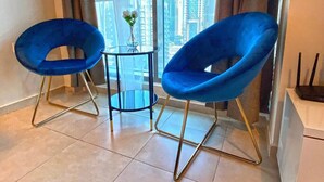 Habitación moderna con dos sillas azules, una mesita y vistas de edificios altos a través de amplias ventanas.
