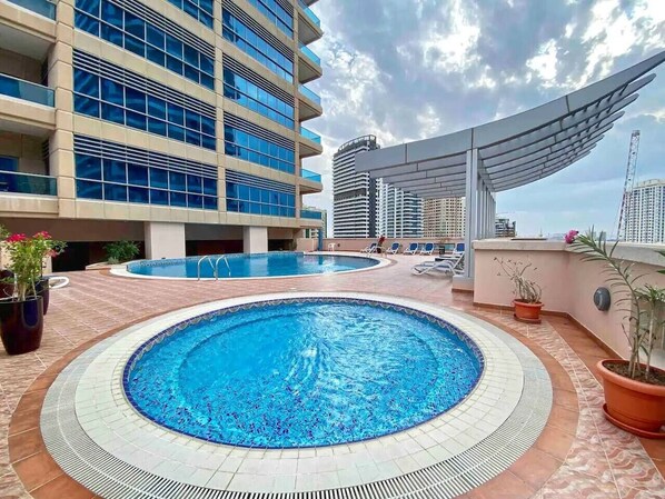 A rooftop pool area with two swimming pools, surrounded by potted plants and lounge chairs.