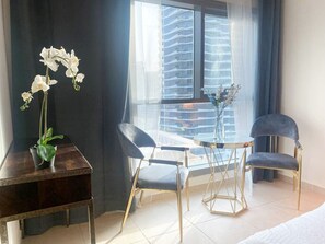A modern hotel room with a glass table, two chairs, a desk with a vase of white flowers, and a view of a cityscape through large windows.