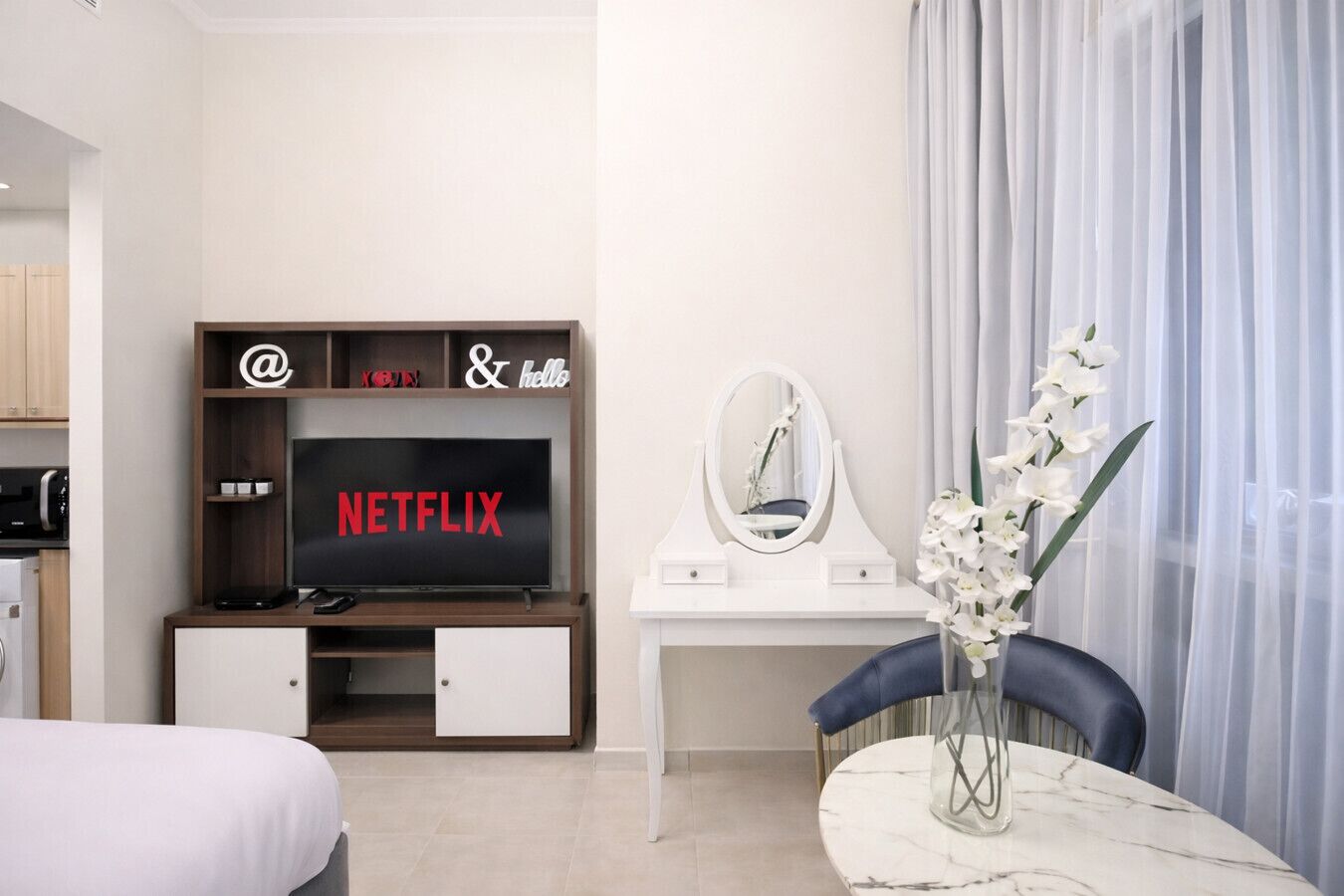 Une chambre d’hôtel avec un lit, un téléviseur affichant le logo de Netflix, un bureau avec un miroir et un vase contenant des fleurs blanches.