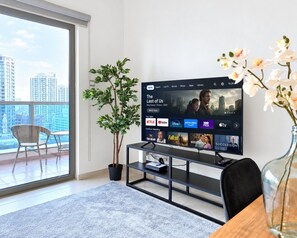 Een moderne woonkamer met een grote flatscreen-tv waarop een streamingdienst wordt weergegeven, een potplant, een eethoek met tafel en stoelen, en een stadsgezicht door een groot raam.