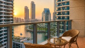 A balcony with wicker chairs and a glass table overlooking a river and cityscape at sunset.