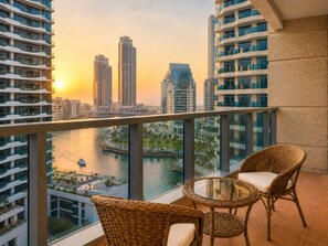A balcony with wicker chairs and a glass table overlooking a river and cityscape at sunset.