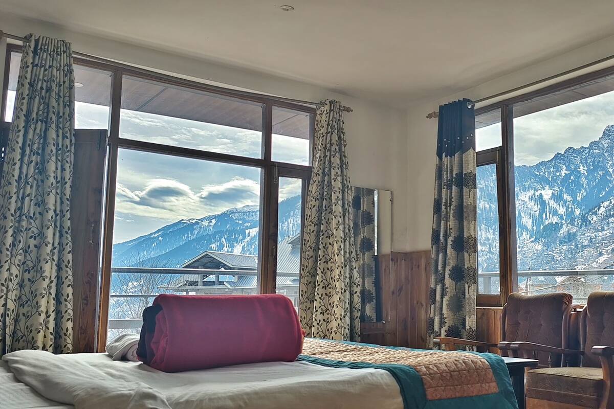 A room with a bed, a desk, and a chair, with large windows offering a mountain view.