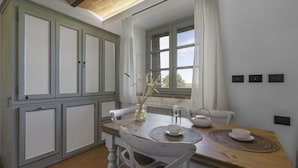 A dining area with a wooden table, white chairs, a vase with dried plants, and a window with curtains. - Nice apartment for 2 guests with pool, WIFI, A/C, TV and pets allowed, close to Siena (Monteriggioni)