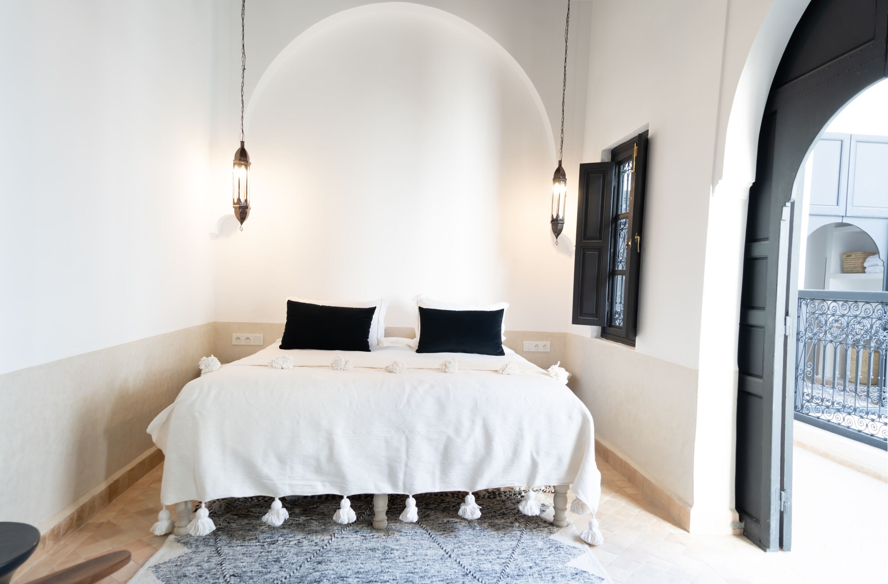 A bedroom with a large bed, two bedside lamps, a black door, and a view of a courtyard.