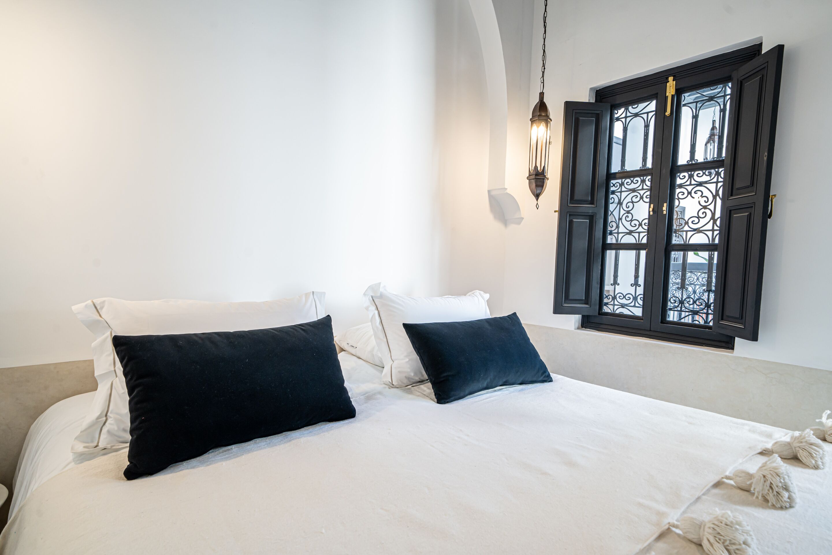 A neatly made bed with white and dark pillows, a hanging light fixture, and a window with decorative iron bars.