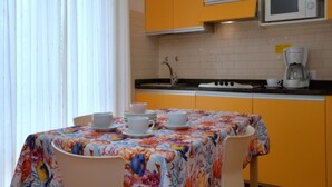 Eine kleine Küche mit gelben Schränken, einer Mikrowelle und einer Kaffeemaschine. Es gibt einen Tisch mit einer blumigen Tischdecke sowie weiße Tassen und Untertassen.