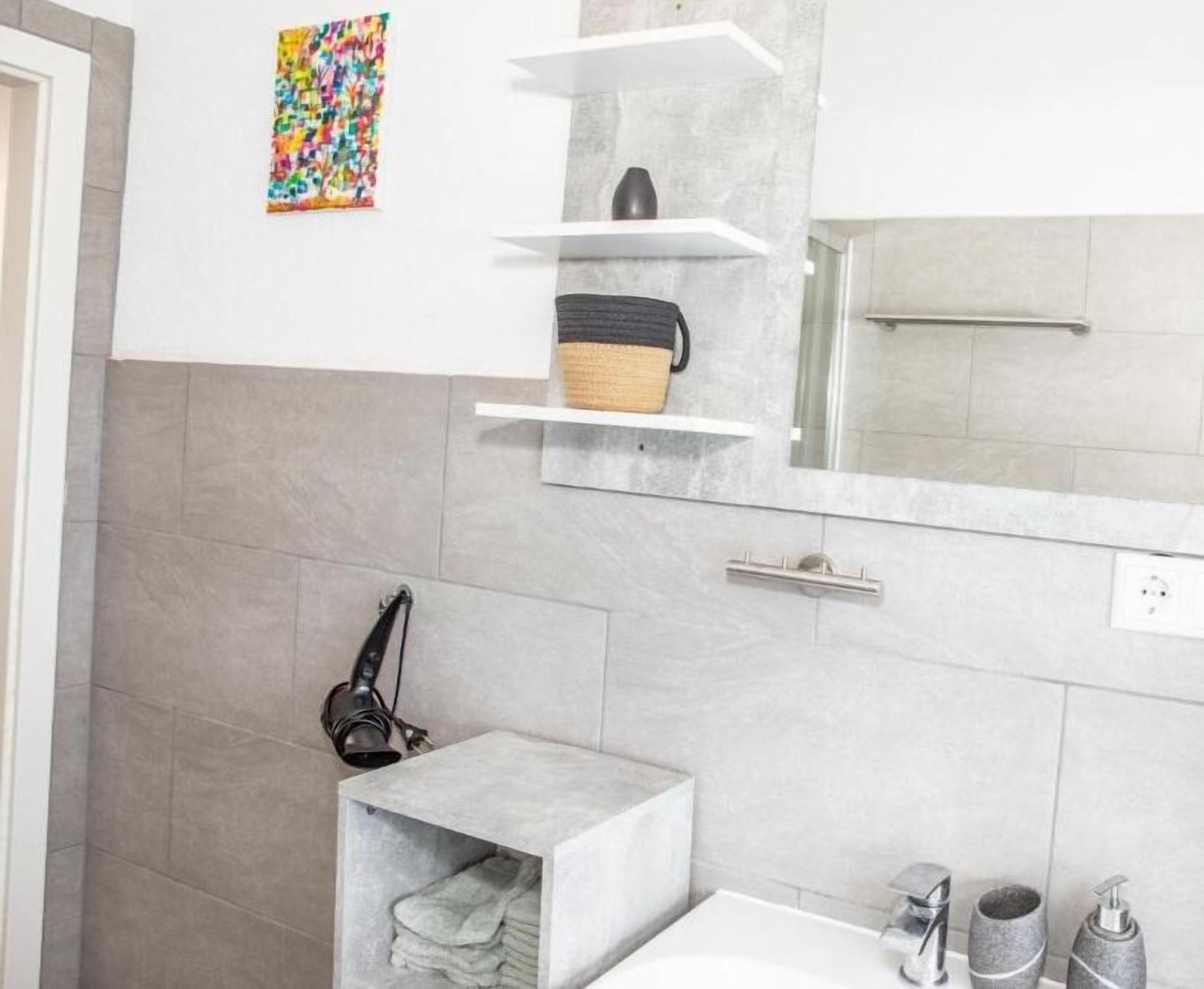 A modern bathroom with grey tiles, a white sink, a mirror, and a shelf with a basket.