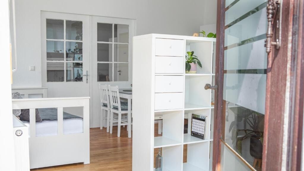 Une pièce compacte et bien éclairée, avec un lit blanc, un petit bureau et une chaise. On y trouve une armoire blanche avec des tiroirs et une étagère sur laquelle est posée une plante en pot.