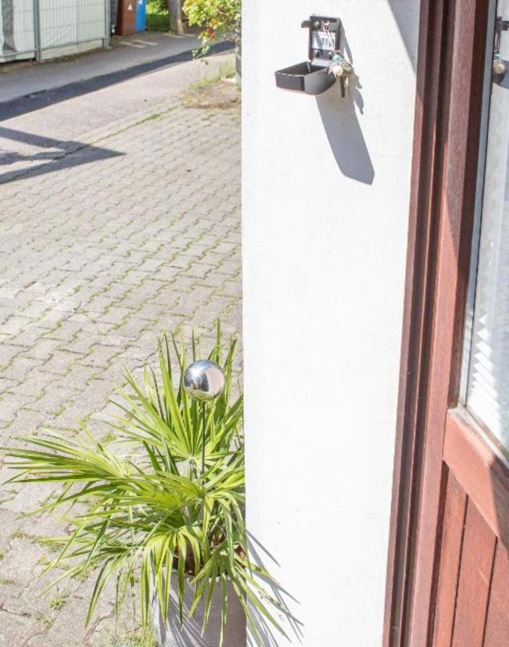 Een potplant met een metalen wereldbolornament op een geplaveid voetpad, naast een gebouw met een witte muur en een houten deur.