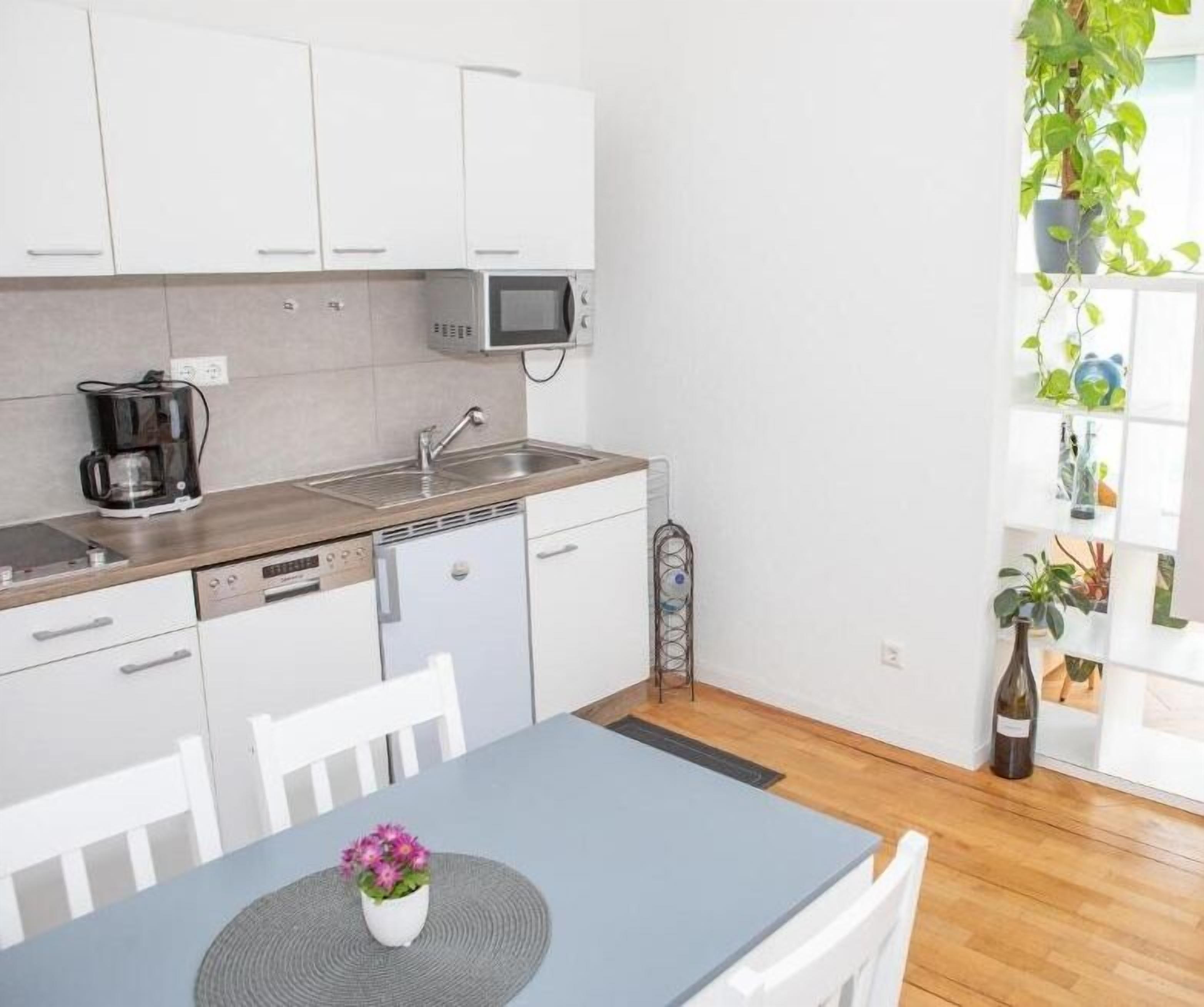 A modern kitchen with white cabinets, a built-in microwave, and a coffee maker. There is a dining table with a vase of flowers.