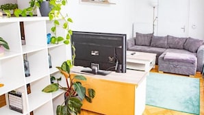 Ein modernes Wohnzimmer mit Holzboden, einem grauen Sofa, einem weißen Schreibtisch mit Computer und mehreren Pflanzen.