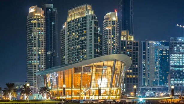 Un edificio moderno in vetro con tetto curvo, circondato da grattacieli dalle finestre illuminate.