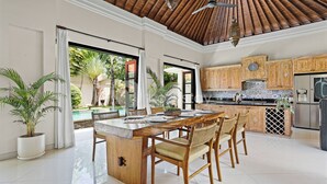 A spacious kitchen with wooden cabinets, a central island, and a view of a swimming pool. - Sombra Villa by Kozystay - Sanur (Denpasar)