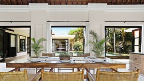 A dining area with a wooden table set for four, surrounded by wicker chairs, with a view of a pool and greenery outside. - Sombra Villa by Kozystay - Sanur (Denpasar)