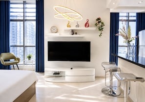 A modern hotel room with a large flat-screen TV, a white console table, a bed, a chair, and a bar area with stools.