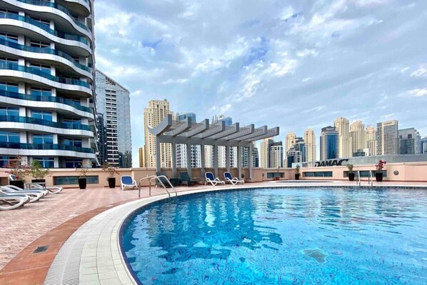 A rooftop swimming pool with lounge chairs, a pergola, and high-rise buildings in the background.